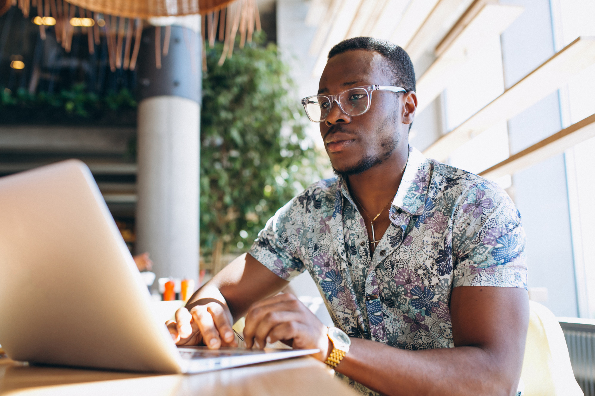A young Black professional wearing glasses and a patterned shirt working intently on a laptop in a bright, modern cafe with natural lighting.