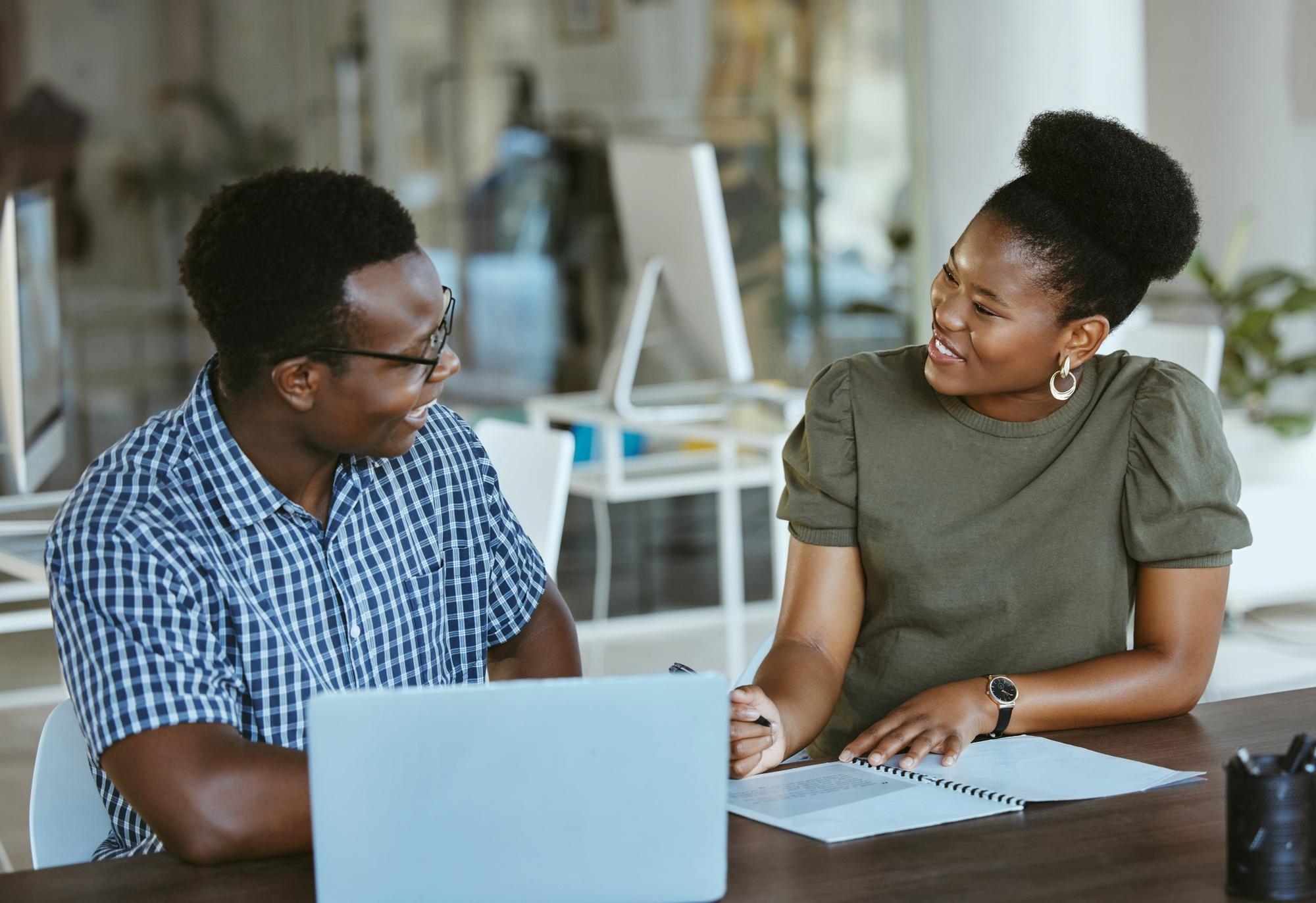 A professional Black man and woman smiling while collaborating on a project with a laptop and notebook in a bright, modern office environment.
