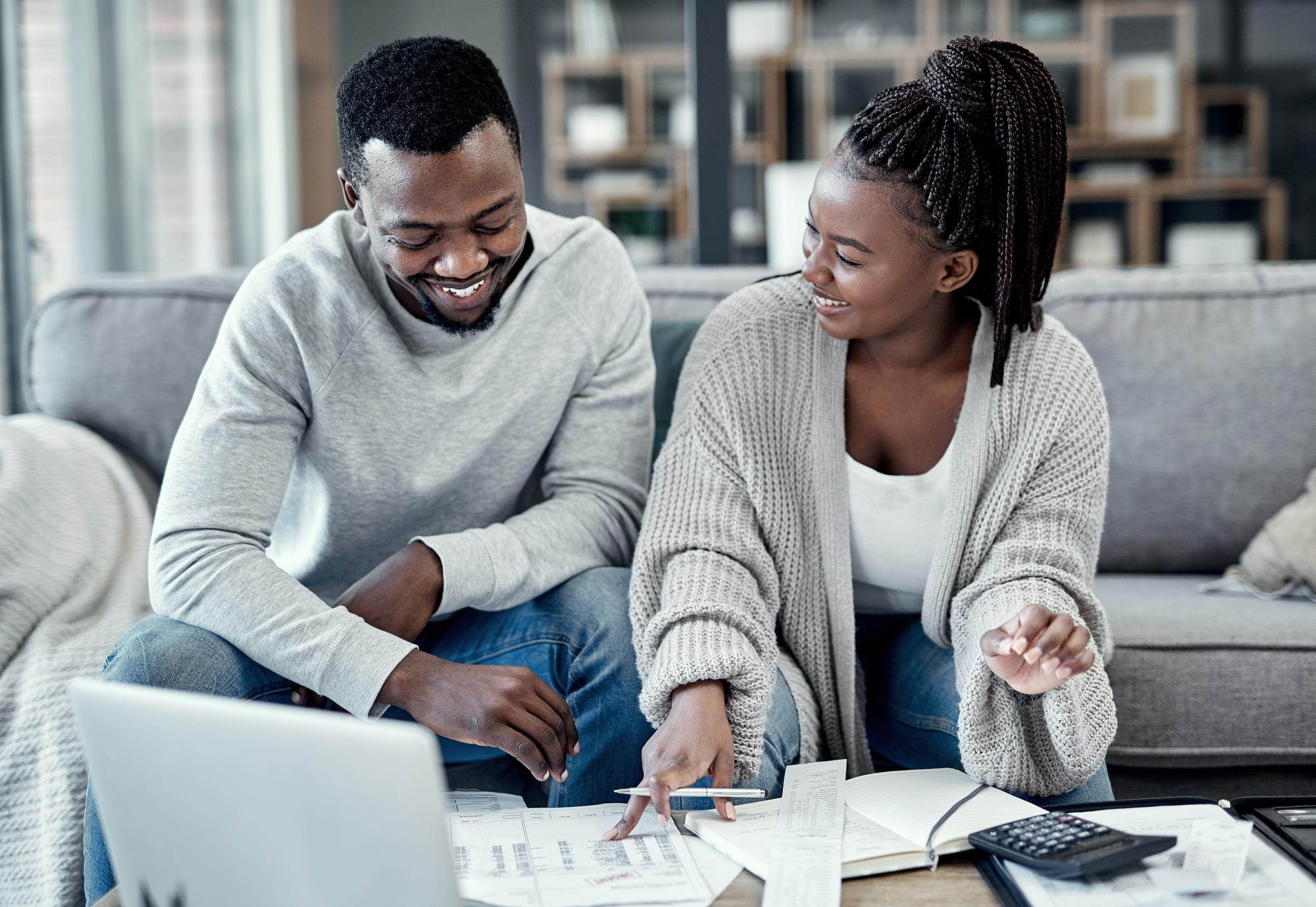 A smiling young Black couple sitting on a sofa and collaborating on household finances with a laptop, calculator, and budget documents.